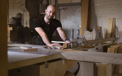 Carpenter in an apron guiding a timber board through a table saw in a workshop
