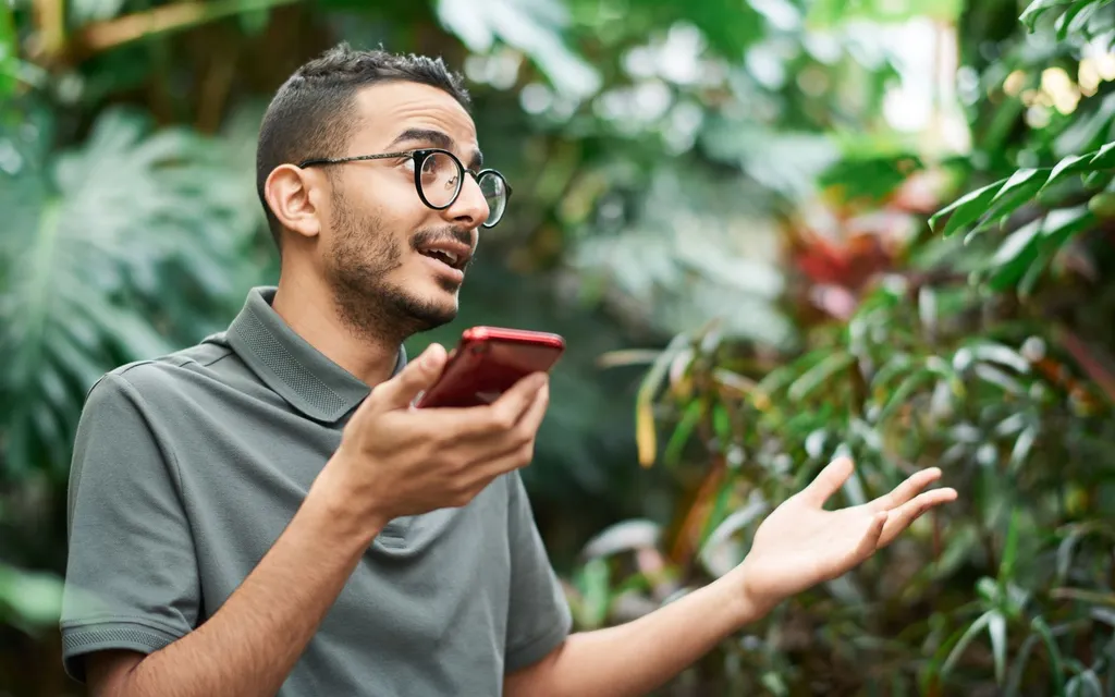 A gardener in glasses taking a call on a phone among lush green plants