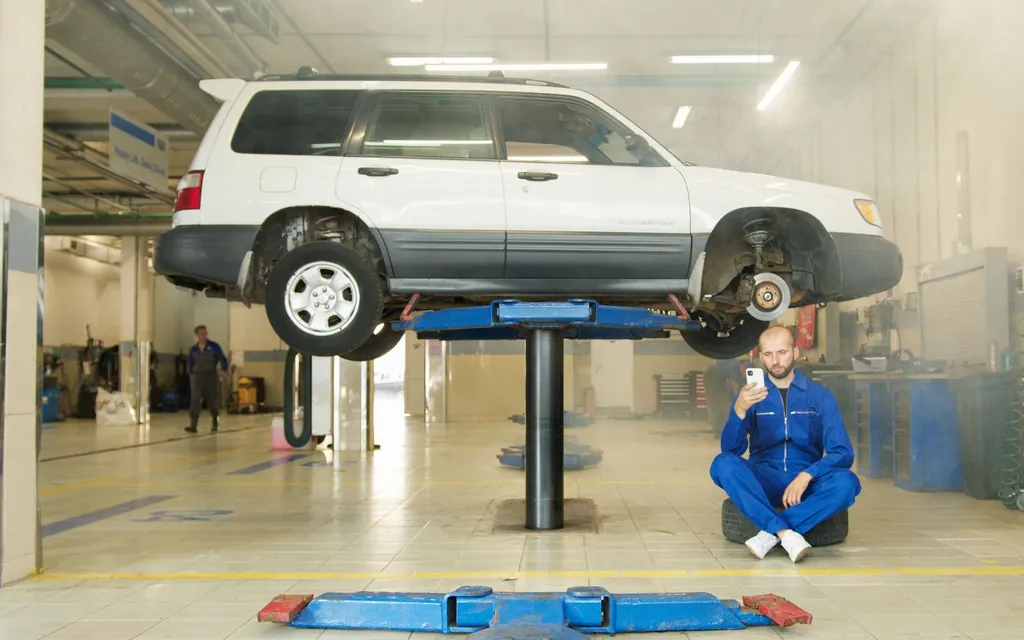 Mechanic in blue overalls sitting on a tyre checking his phone, a car raised on the ramp behind him