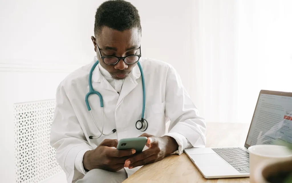 A doctor in a white coat and stethoscope checking a smartphone at the practice desk