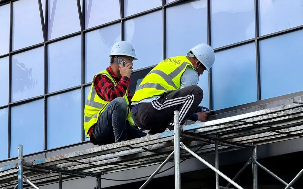 Two roofers in hi-vis and hard hats on scaffolding, one taking a call on a phone